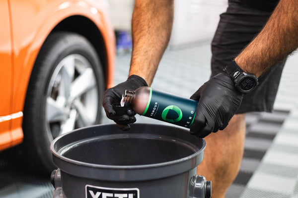A closeup of a person wearing black rubber gloves pouring Armour Detail Hero into a black bottle cap over a black bucket with an orange car in the background.