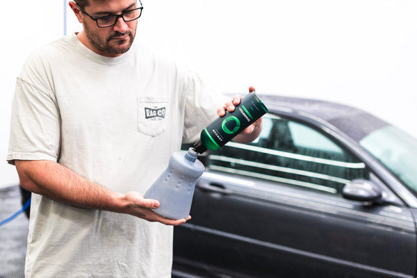A person pouring Armour Shampoo+ into a foam cannon in front of a grey car.