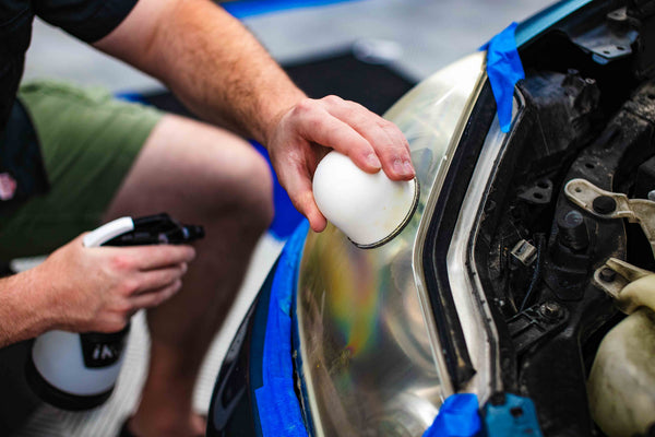 A person holding the IK Mini Sprayer in the right and and the Headlight kit hand block in the left hand with a blue car in the background.