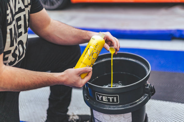 A person pouring a bottle cap full of Opti-Coat No Rinse into a black bucket full of water and bubbles.