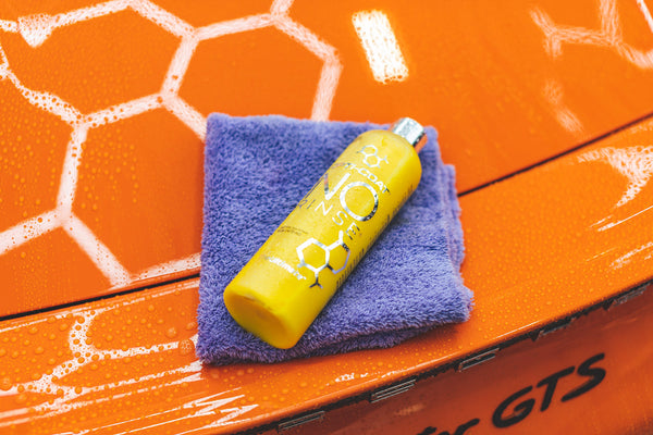 A bottle of Opti-Coat laying on a periwinkle towel on the back of an orange Porsche.