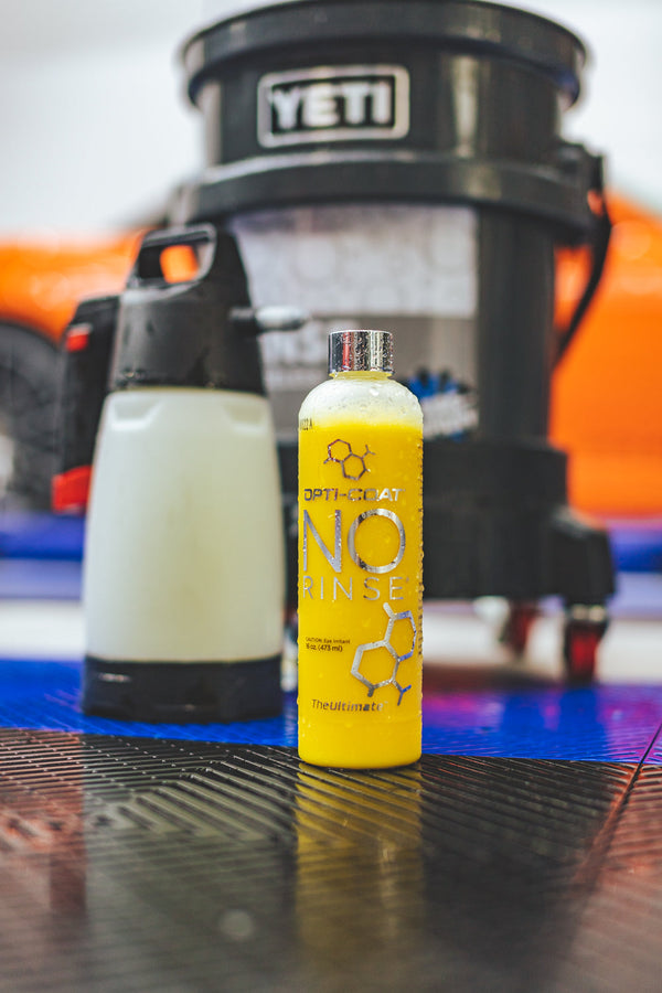A bottle of Opti-Coat No Rinse in front of a black bucket and an IK Sprayer inside of an autobody shop..