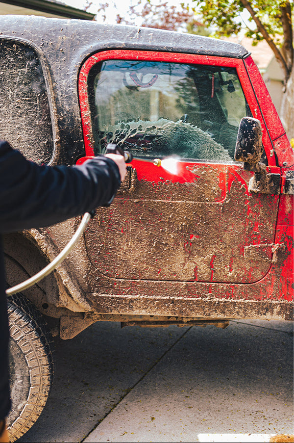 A closeup of a person spraying a muddy red jeep with a power washer.