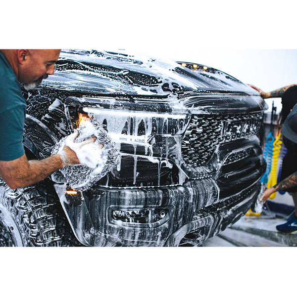 The front of a black truck covered in foam while to people scrub it with long fiber towels.