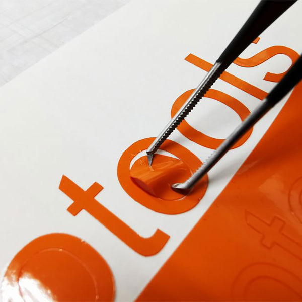 Close-up of a pair of Yellotool Sign tweezers carefully peeling off a section of orange vinyl from a white surface, revealing part of a letter in the word 'tools' beneath.